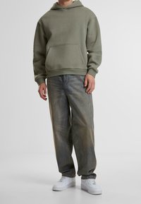 Felpa con cappuccio verde oliva con tasca frontale, abbinata a jeans blu slavati a vita bassa e sneakers bianche. Tessuto dalla texture liscia.