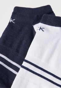 Δύο διπλωμένες κάλτσες αστραγάλου, μία σε ναυτικό μπλε και μία λευκή, κάθε μία με δύο οριζόντιες ρίγες κοντά στο πάνω μέρος της μανσέτας.