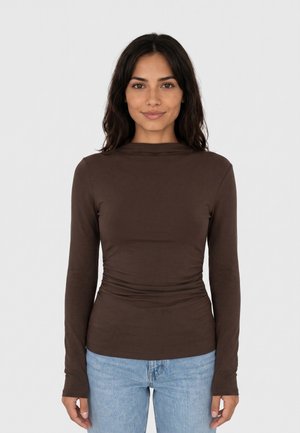 Giovane donna con lunghi capelli scuri che indossa un top marrone a maniche lunghe e jeans azzurri chiari, in piedi davanti a uno sfondo chiaro e uniforme, rivolta verso il davanti.