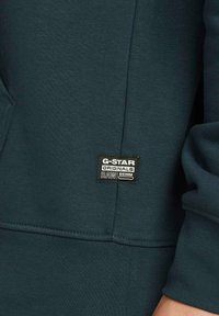 Ciemnoniebieska bluza z kapturem wykonana z teksturowanej tkaniny, z kieszenią kangurzą oraz czarną metką z białym napisem "G-STAR ORIGINALS RAW DENIM."