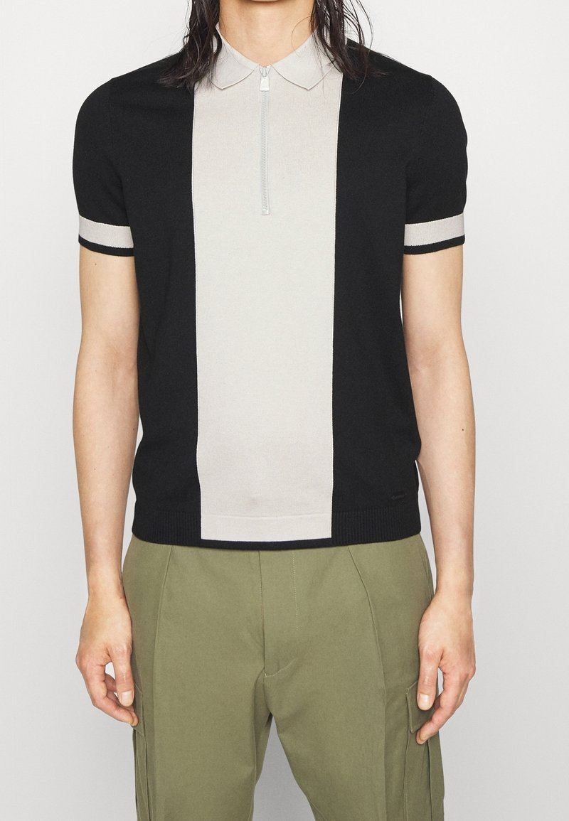 Personne portant un polo à manches courtes noir et beige avec un panneau central beige et un col zippé, associé à un pantalon vert olive.