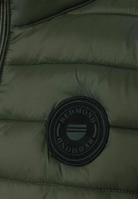Grüne Pufferjacke aus Stoff mit horizontalen Steppabschnitten. Verfügt über einen runden schwarzen Aufnäher mit dem Text 'REDMOND' und einem kreisförmigen Design.