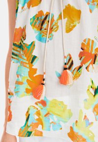 Weißes Oberteil mit bunten Blattmustern in Orange, Blau und Grün. Enthält einen Kordelzug mit Quasten in Orange und schwarzen Akzenten.