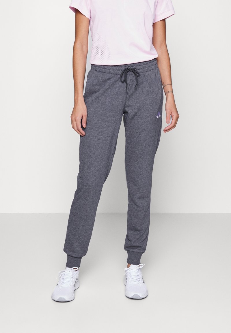 Pantalons de survêtement en coton gris avec taille élastique et cordon de serrage. Comprend des poignets côtelés. Portés avec un t-shirt rose clair et des chaussures de sport blanches.