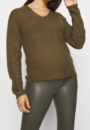 Vrouw die een olijfgroene geribbelde gebreide trui en donkergrijze leren legging draagt, met creolen, poserend tegen een effen achtergrond.