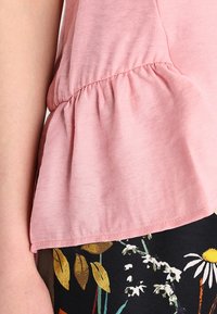Světle růžový peplum top s jemnou texturou, nabírané švy v pase, spárovaný s černými kalhotami s květinovým vzorem, které mají žluté a bílé květy.