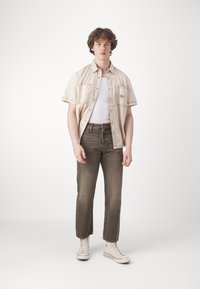 Lys beigefarget kortermet skjorte over en hvit tanktopp, brune rette jeans, og hvite høye sneakers på en ensfarget bakgrunn.