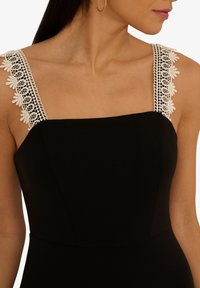 Robe noire avec un décolleté carré et des bretelles en dentelle décoratives avec des motifs floraux. Le tissu lisse crée une silhouette ajustée.