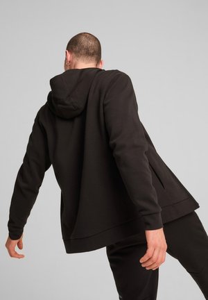 Mężczyzna pochyla się do tyłu, ubrany w jednolitą czarną bluzę z kapturem i czarne spodnie na szarym tle, głowa odwrócona od kamery.