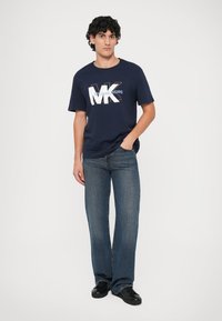 Camiseta de algodón azul marino con gran logo blanco "MK" y texto "MICHAEL KORS", combinada con jeans de pierna recta y zapatos negros.