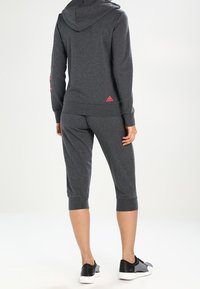 Szara bluza z kapturem z różowymi akcentami i logo, w połączeniu z pasującymi spodniami typu capri i czarnymi butami sportowymi. Widoczna gładka struktura tkaniny.