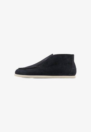 Schwarzer Veloursleder-Slipper mit weißer flacher Sohle und minimaler Naht, seitlich auf weißem Hintergrund.