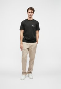 Μαύρο βαμβακερό T-shirt με λογότυπο στο στήθος, συνδυασμένο με μπεζ παντελόνι και λευκά αθλητικά παπούτσια. Απλός, casual σχεδιασμός με χαλαρή γραμμή.