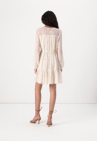 Robe crème à manches courtes avec une jupe à volants, tissu transparent, motif à pois dorés et taille à cordon. Associée à des talons beiges à lanières.