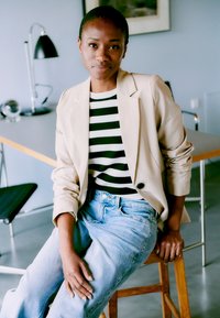 Frau in beigem Blazer, schwarz-weiß gestreiftem Hemd und hellblauen Jeans, die auf einem Holzschemel in modernem Büroumfeld sitzt.