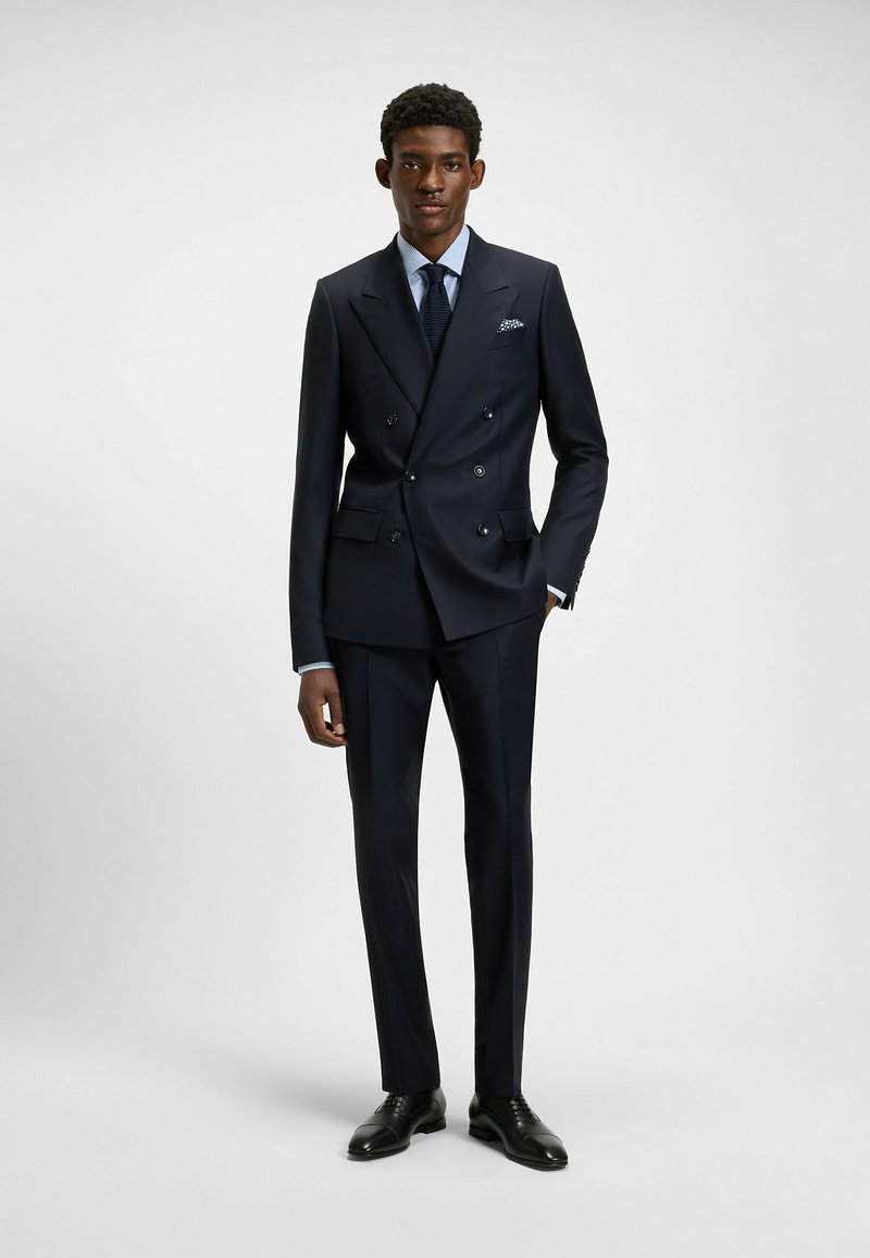 Navy double-breasted suit with notch lapels, matching trousers, a light blue shirt, tied with a dark tie, and black dress shoes.