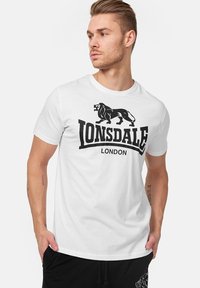 Fehér pamut póló fekete oroszlán logóval és "LONSDALE LONDON" felirattal. Rövid ujjú és kerek nyakkivágású, laza fazonú.