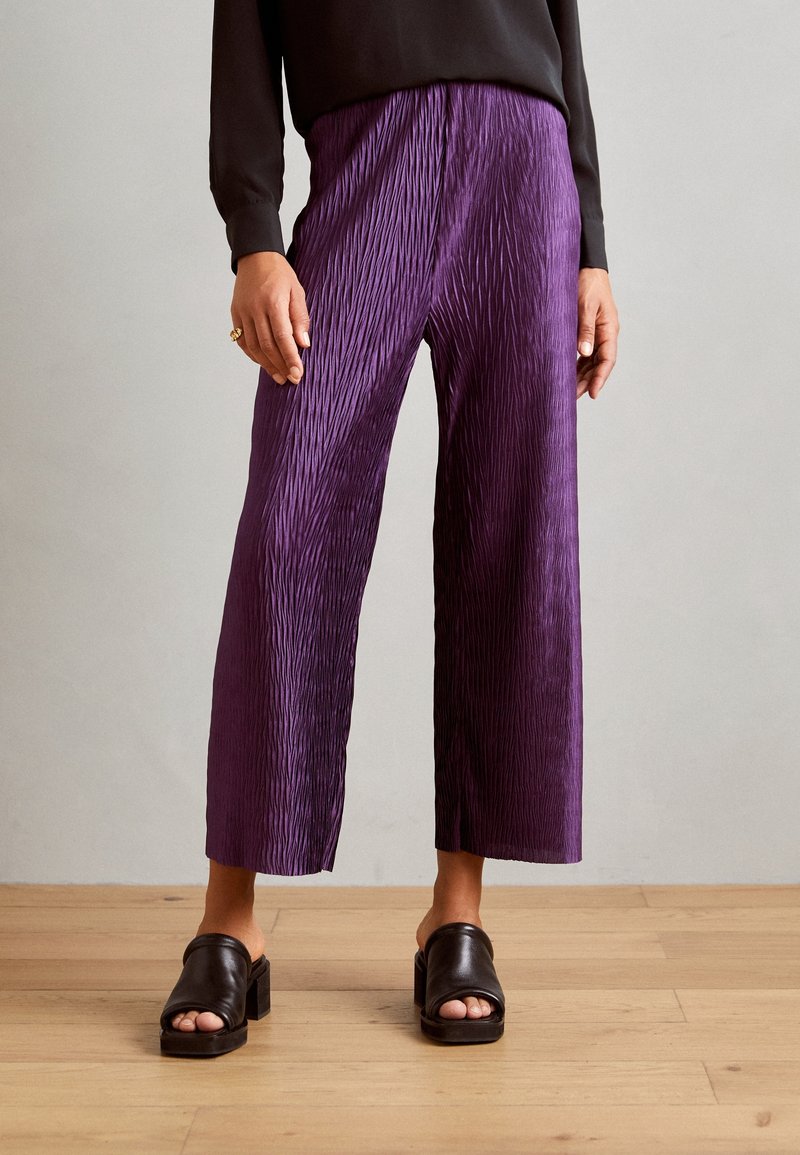 Femme portant un pantalon large texturé violet, un haut noir à manches longues, des sandales à talons blocs noires ouvertes à l'avant, debout sur un sol en bois.