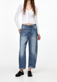 Světle modré džíny s širokým střihem, s vybledlými detaily a ohrnutými nohavicemi, spárované s bílým žebrovaným zipovým topem a černými loafery.