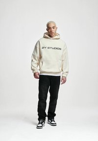 Felpa color crema con tasca frontale e testo nero "2Y STUDIOS", abbinata a jeans neri e sneaker bianco e nere.