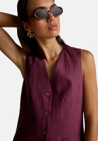 Kobieta w bezrękawnikowej kamizelce w kolorze śliwkowym z guzikami, w okularach przeciwsłonecznych w kształcie owalnym z żółwią skorupą oraz złotych kolczykach w kształcie gałązek, pozująca z uniesioną jedną ręką.