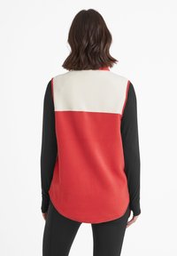 Red and cream color-blocked fleece vest with a round hem. Soft texture, sleeveless design over a black long-sleeve shirt.