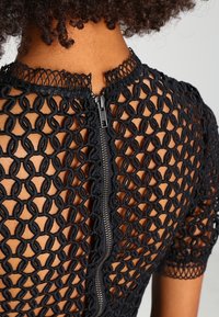 Černý krajkový top s geometrickým výřezem, na zádech malý zip. Krátké rukávy s vlnkovanými okraji.