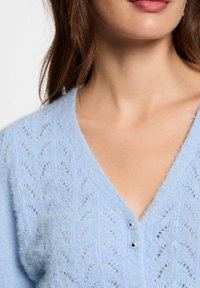 Cárdigan de punto azul claro con escote en V, que presenta un patrón de encaje, tejido texturizado y dos pequeños botones plateados en el cuello.