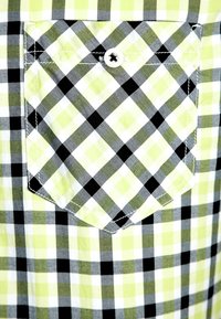 Poche à boutons sur une chemise en tissu à carreaux verts, noirs et blancs avec un motif à carreaux diagonal sur la poche.