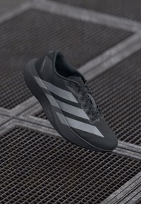 Tmavě šedá běžecká obuv Adidas se světle šedými pruhy, s zavázanými tkaničkami, visící nad kovovou mřížkovou plochou.