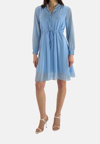 Robe bleu clair avec de longues manches transparentes, un bouton à l'avant, une taille à cordon et un ourlet évasé. Portée avec des talons blancs à lanières.