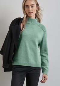 Mintgrön tröja med hög ribbad krage, prydd med dekorativa knappar på axeln samt ribbade ärmslut och nederkant. Bärs med svarta jeans.