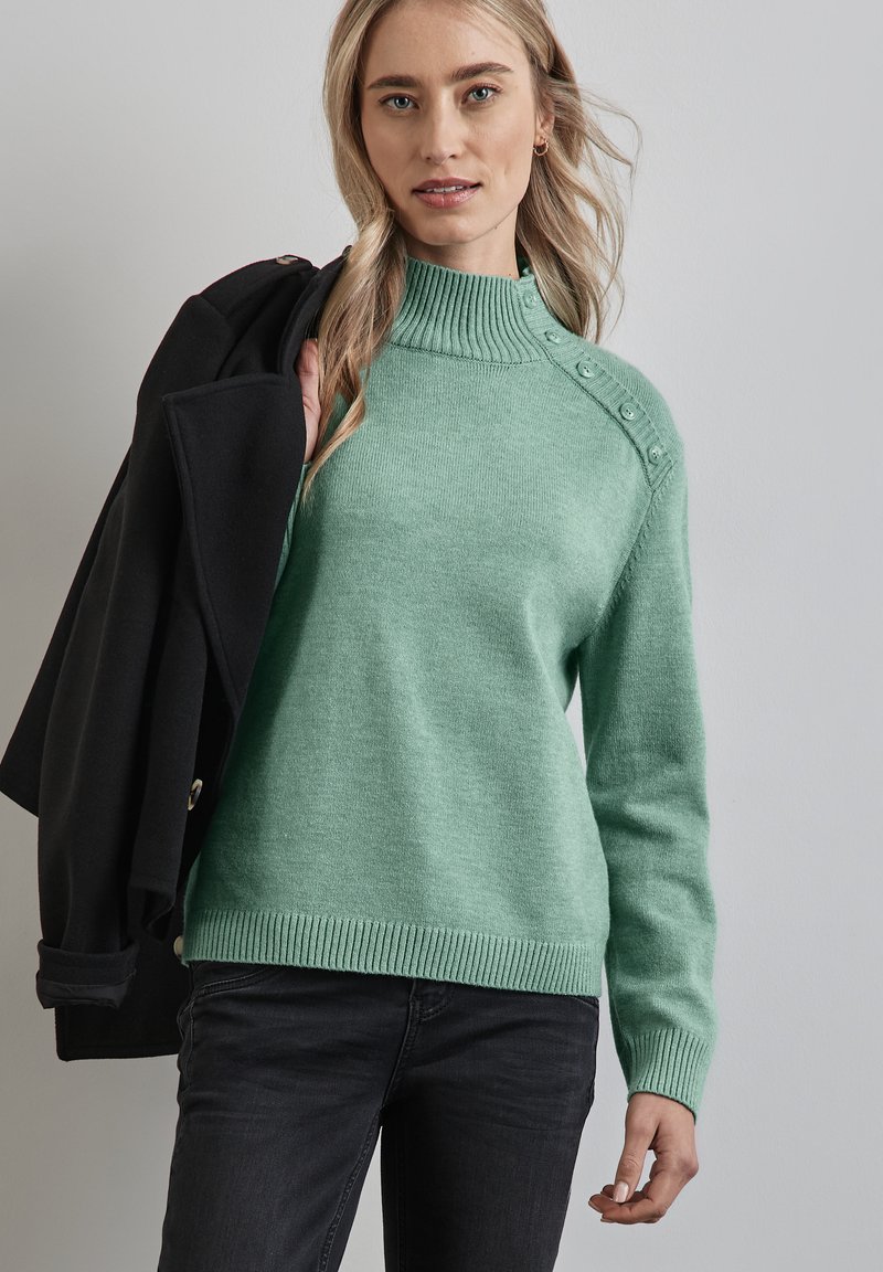 Mintgrön tröja med hög ribbad krage, prydd med dekorativa knappar på axeln samt ribbade ärmslut och nederkant. Bärs med svarta jeans.