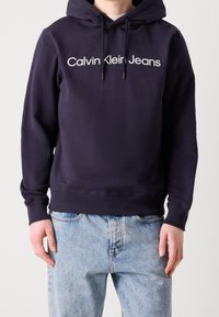 Marinblå hoodie i mjukt tyg med en framficka, vit "Calvin Klein Jeans"-logotyp, dragsko i huvan och ribbat avslut.