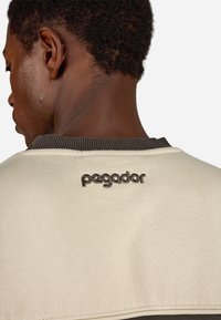 Off-white mikina s tmavým žebrovaným límcem. Na zadní straně je vyšité slovo "pegador" tmavou nití. Hladká textura látky.