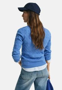 Blue cable-knit sweater with a ribbed hem, layered over a white and blue striped shirt, paired with light denim and a navy cap.