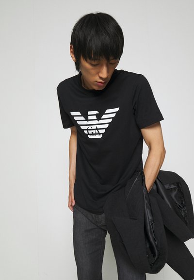 Black cotton t-shirt featuring a white eagle logo design on the front. The model wears dark jeans and holds a black jacket.