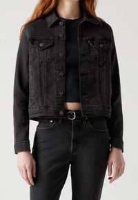 Chaqueta de mezclilla negra con un diseño corto, cierres de botón, dos bolsillos en el pecho y una textura suave. Combinada con unos vaqueros negros de cintura alta.