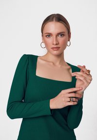Zielona, prążkowana bluzka z długim rękawem i kwadratowym dekoltem. Modelka nosi duże kolczyki w kształcie kół oraz warstwowe pierścionki na palcach. Gładka tekstura skóry.
