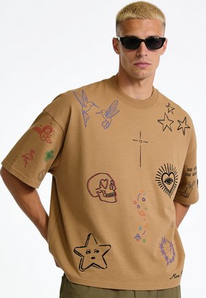 Homme blond portant des lunettes de soleil noires et un t-shirt beige oversize avec des imprimés colorés de style tatouage d'oiseaux, crâne, étoiles et cœurs.