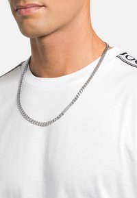 Collar de cadena plateada con eslabones entrelazados y un acento rectangular plano, llevado sobre una camisa blanca sencilla.