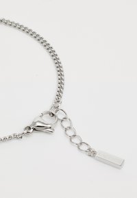 Silver chain bracelet with interlocking links, featuring a lobster clasp and a rectangular charm inscribed with "LACOSTE." Smooth texture.