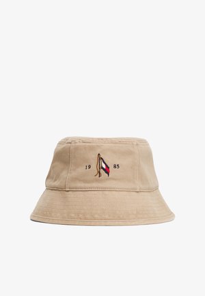 Beżowy kapelusz typu bucket wykonany z bawełny, z płaskim rondem. Posiada haftowane logo w czerwonych, białych i niebieskich akcentach oraz liczby "1985".