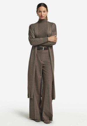 Donna in piedi con le braccia incrociate, indossa un cardigan lungo marrone, un maglione a collo alto, pantaloni a gamba larga e una cintura scura su sfondo bianco.