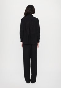 Zwarte, lange mouwen shirt met een kraag en knoopmanchetten, gedragen over losse zwarte broek, gezien van achteren op een lichte achtergrond.