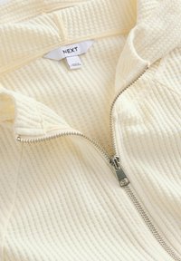 Sudadera con cremallera color crema, con un patrón texturizado y acanalado, cremallera plateada y una etiqueta que dice "NEXT" dentro del cuello.