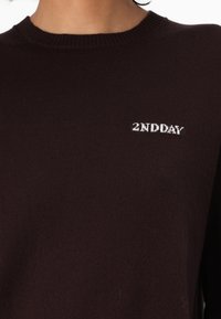 Nahaufnahme eines dunkelbraunen Strickpullovers mit weißem gesticktem Text "2ND DAY" auf der oberen Brust nahe dem Halsausschnitt.