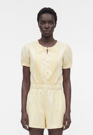 Kobieta ubrana w żółto-biały kraciasty kombinezon z krótkim rękawem, z guzikami i wiązaniem przy dekolcie, stojąca na jednolitym białym tle.