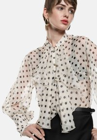 Blusa cremisi trasparente con pois neri, parte anteriore con ruches, collo alto e maniche lunghe a palloncino; chiusura a bottoni discreta al colletto.
