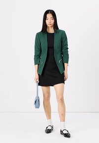 Zelený blazer s měkkými kapsami, spárovaný s černými bezramínkovými šaty. Modelka drží světle modrou kabelku a má na sobě bílé ponožky a černé boty.